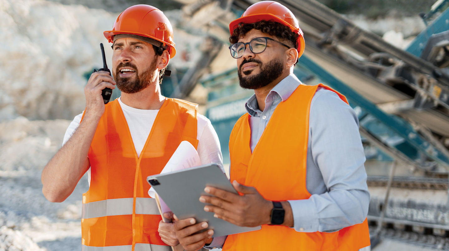 Chef de chantier en casque rouge montrant l'écran d'une tablette durcie à deux ouvriers en casques blanc et jaune sur chantier avec containers en arrière-plan